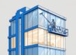 A window cleaner on a suspended platform washes an office building’s glass exterior.