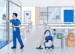 Cleaner wiping a glass door in a bright, empty office with boxes and cleaning supplies.